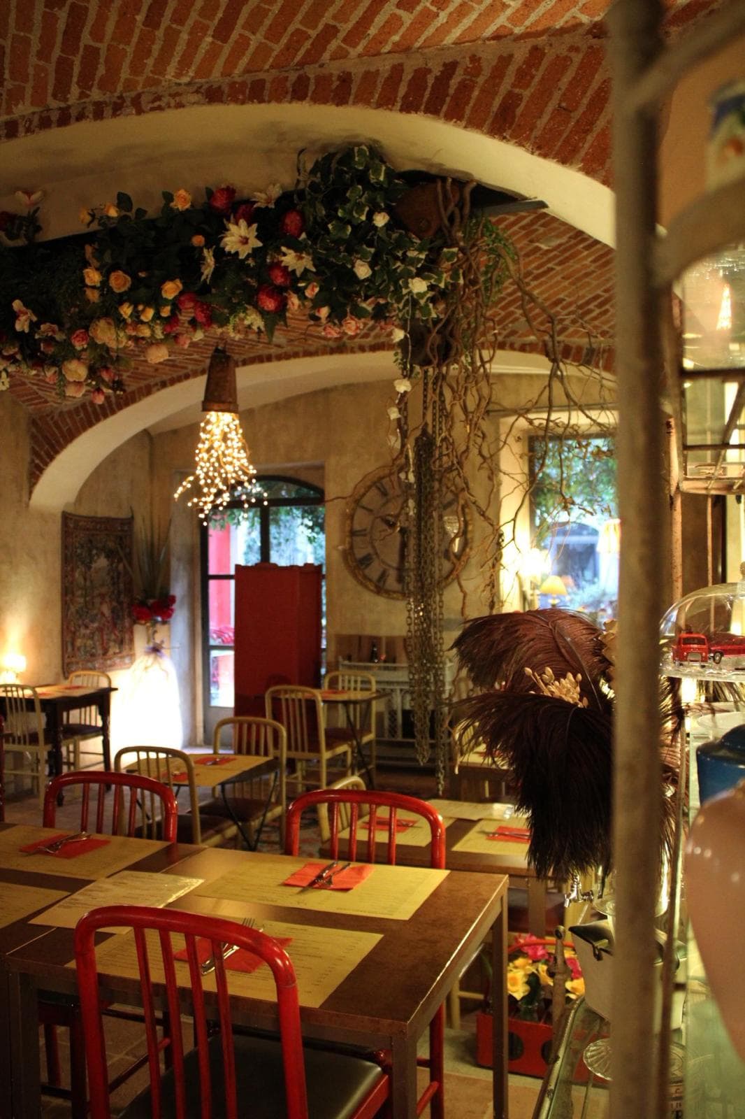La sala sotto la volta di mattoni: tavoli con le carte, sedie rosse e fiori appesi all'arco.